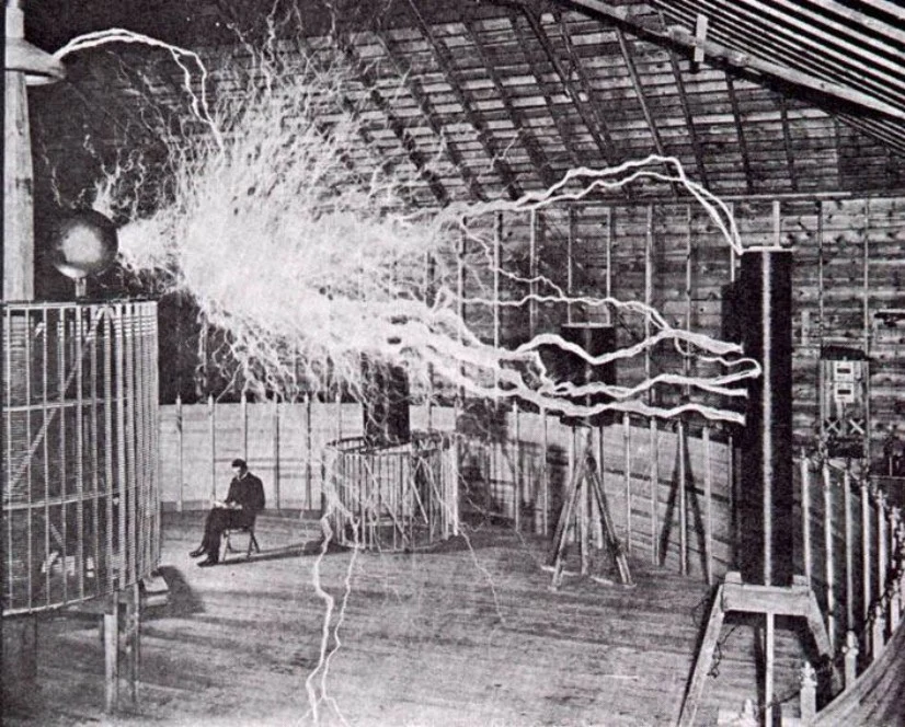 Nikola Tesla sitzt ruhig lesend in seinem Colorado Springs Labor, während riesige künstliche Blitze von seinem Resonatortransformator durch die Luft zucken — Doppelbelichtung, 1899